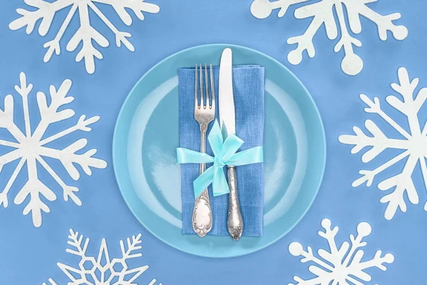 Vue surélevée de la fourchette et du couteau enveloppés par un ruban festif sur une assiette entourée de flocons de neige isolés sur du bleu — Photo de stock