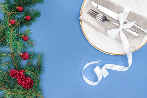 Flat lay with fork and knife wrapped by ribbon on plate near evergreen branches with christmas balls and red berries isolated on blue — Stock Photo