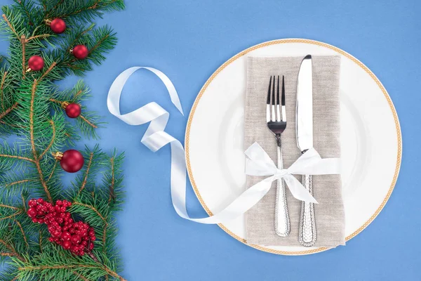 Верхний вид вилки и ножа, завернутые лентой на тарелке рядом с вечнозелеными ветвями с рождественскими шариками и красными ягодами, изолированными на голубом — стоковое фото