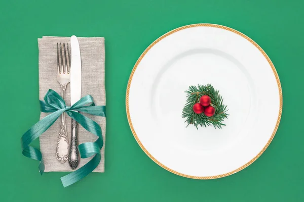 Flat lay with evergreen branch and chiristmas balls on plate near fork with knife wrapped by ribbon isolated on green — Stock Photo