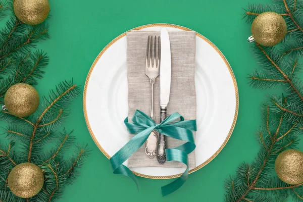 Blick von oben auf Gabel und Messer, umwickelt von festlicher Schleife auf Teller, umgeben von immergrünen Baumzweigen mit Weihnachtskugeln auf grünem Grund — Stockfoto