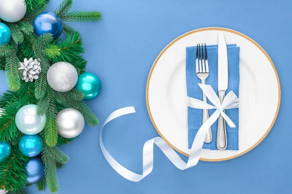 Вид сверху на вилку и нож, завернутые праздничной лентой на тарелке возле вечнозеленых ветвей деревьев с рождественскими шариками, изолированными на голубом — стоковое фото