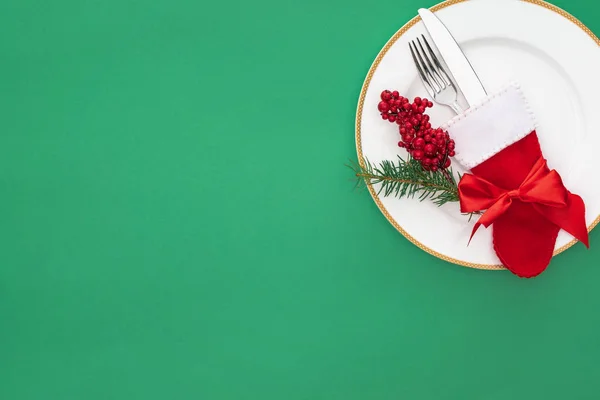 Vista sopraelevata di bacche rosse, ramo d'albero sempreverde e forchetta con coltello in calza natalizia su piatto isolato su verde — Stock Photo