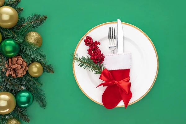 Flat lay with red berries and fork with knife in christmas sock on plate near evergreen branches with christmas balls isolated on green — Stock Photo