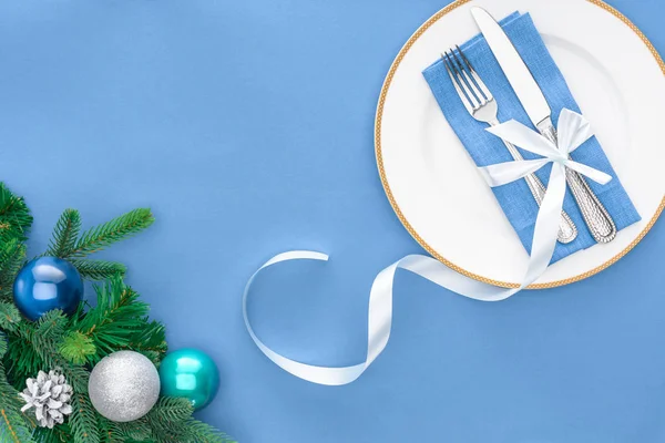 View from above of fork and knife wrapped by festive ribbon on plate near evergreen tree branches with christmas balls isolated on blue — Stock Photo