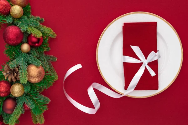 Vista superior de la servilleta envuelta por cinta festiva en el plato cerca de ramas de árboles de hoja perenne con bolas de Navidad aisladas en rojo - foto de stock