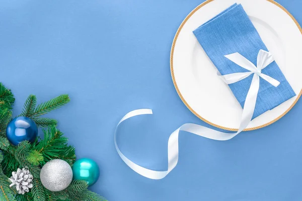 Flat lay with napkin wrapped by festive ribbon on plate near evergreen tree branches with christmas balls isolated on blue — Stock Photo