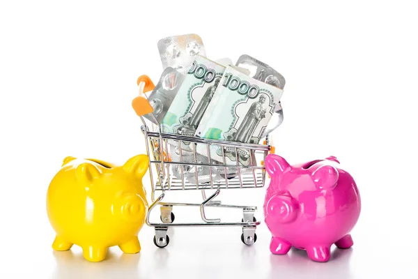 Trolley cart with russian cash money and empty pills packages near pink piggy bank and colorful pills isolated on white — Stock Photo