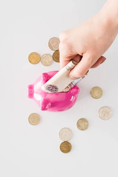 Cropped image of woman putting russian banknote into pink piggy bank surrounded by coins on white surface — Stock Photo