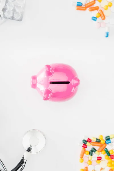 Cama plana con alcancía rosa rodeado de coloridas píldoras varias, estetoscopio y paquetes de pastillas vacías en la superficie blanca - foto de stock