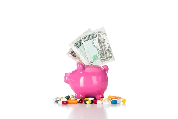 Pink piggy bank with russian banknotes surrounded by various colorful pills isolated on white — Stock Photo