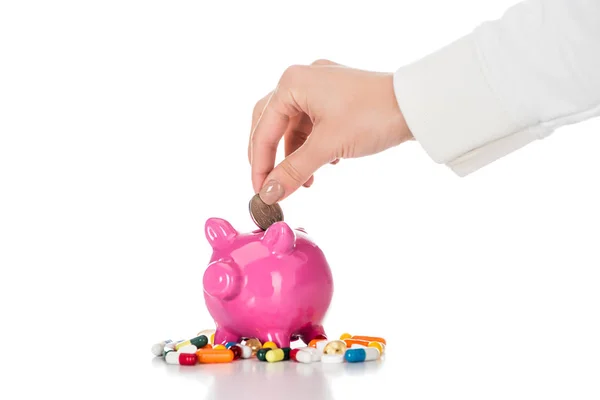 Cropped image of woman putting coin into pink piggy bank surrounded by colorful pills isolated on white — Stock Photo