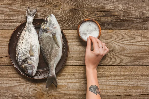 Cropped image of tattooed woman salting uncooked fish on wooden table — Stock Photo