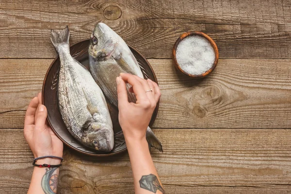 Partial view of tattooed woman salting uncooked fish on wooden table — Stock Photo