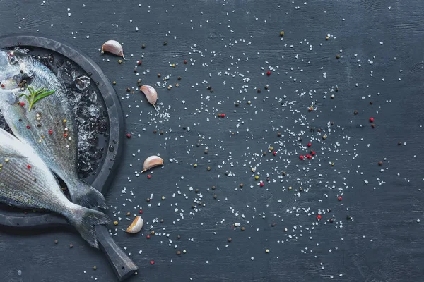 Elevated view of raw fish wit rosemary in tray on black table with garlic — Stock Photo