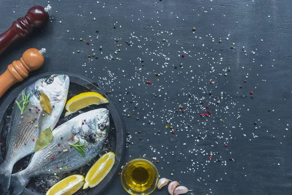 Vista superior de pescado crudo con ingredientes en bandeja sobre mesa negra cubierta de sal y pimienta - foto de stock