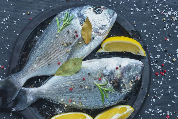 Flach mit rohem Fisch mit Zitrone, Lorbeerblättern und Rosmarin in Tablett auf schwarzem Tisch mit Salz und Pfeffer bedeckt — Stockfoto
