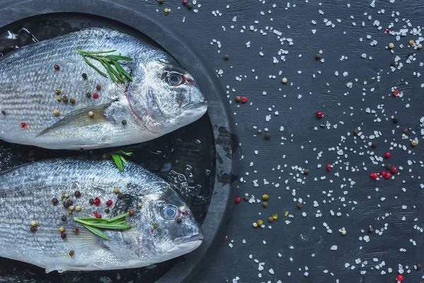 Elevated view of raw fish with rosemary in tray on black table covered by salt and pepper — Stock Photo
