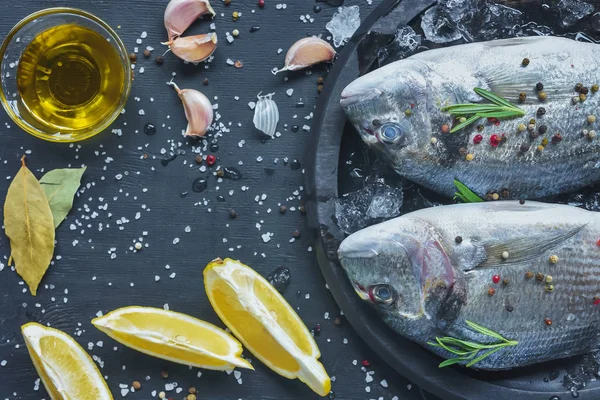 Elevated view of raw fish covered by pepper and rosemary in tray on black table — Stock Photo