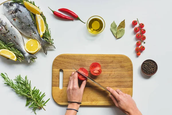 Imagen recortada de mujer cortando tomate por cuchillo en tablero de madera cerca de pescado crudo en la mesa - foto de stock