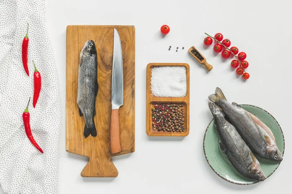 Vista elevada del pescado sin cocer en una tabla de madera cerca de los ingredientes en la mesa blanca - foto de stock