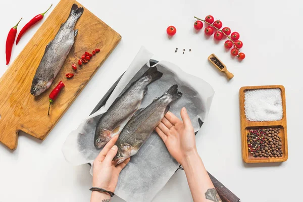 Частичный вид женщины, положившей сырую рыбу в лоток с выпечкой бумаги на белый стол с ингредиентами — стоковое фото