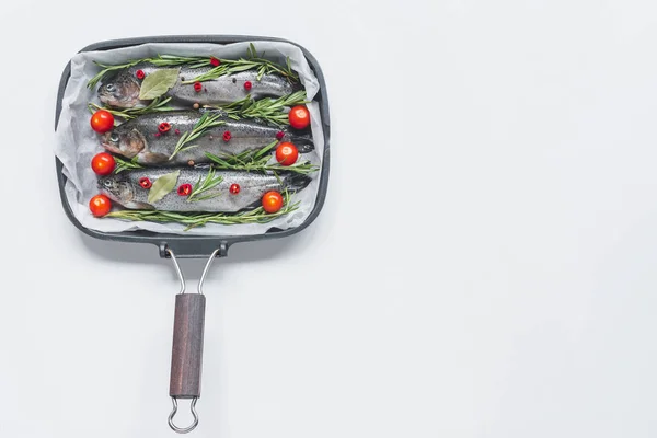 Vue du dessus du poisson avec romarin, feuilles de laurier et tomates cerises dans une plaque avec papier cuisson sur table blanche — Photo de stock