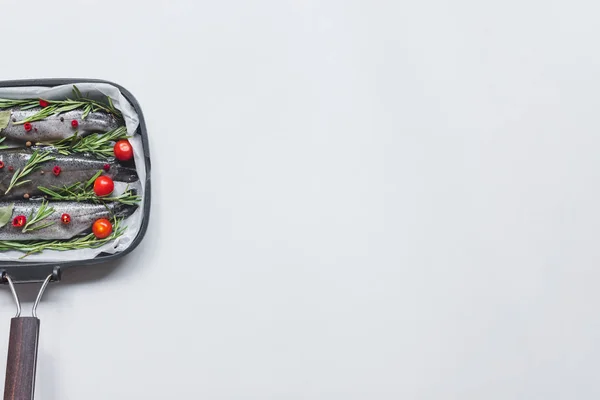 Vista elevada de peixes com alecrim, folhas de louro e tomates cereja em bandeja com papel manteiga sobre mesa branca — Fotografia de Stock