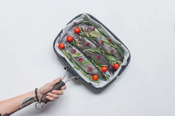 Abgeschnittenes Bild Frau hält Tablett mit Fisch mit Rosmarin, Lorbeerblättern und Kirschtomaten über weißem Tisch — Stockfoto