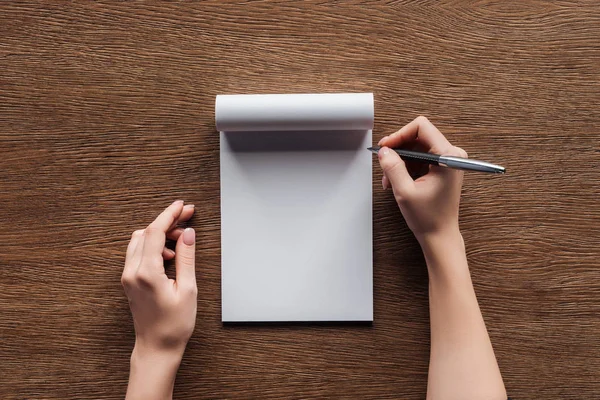 Vista cortada da pessoa segurando caneta sobre notebook em branco no fundo de madeira — Fotografia de Stock