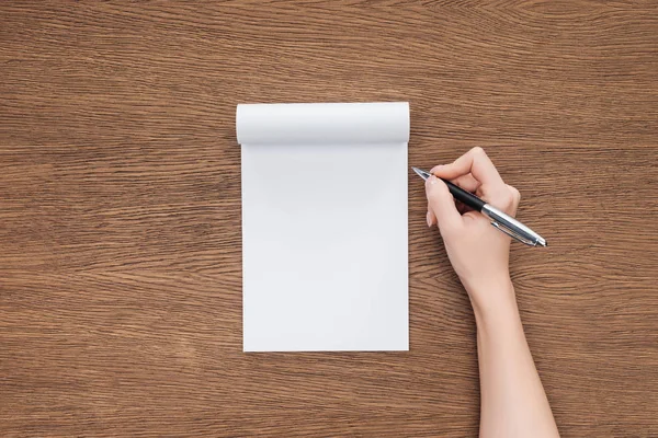 Vista cortada da pessoa segurando caneta sobre notebook em branco no fundo de madeira — Fotografia de Stock