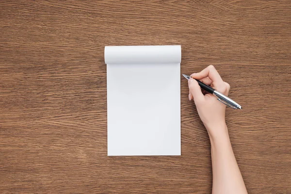 Vista recortada de la persona que sostiene la pluma sobre el cuaderno en blanco sobre fondo de madera - foto de stock