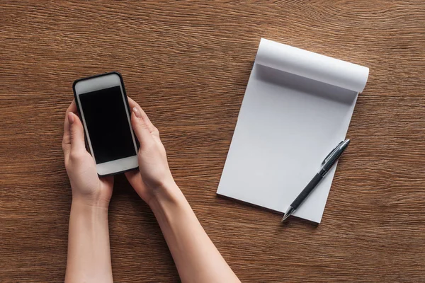 Vista parziale di persona che tiene smartphone con schermo bianco, penna, notebook con spazio su sfondo in legno — Foto stock