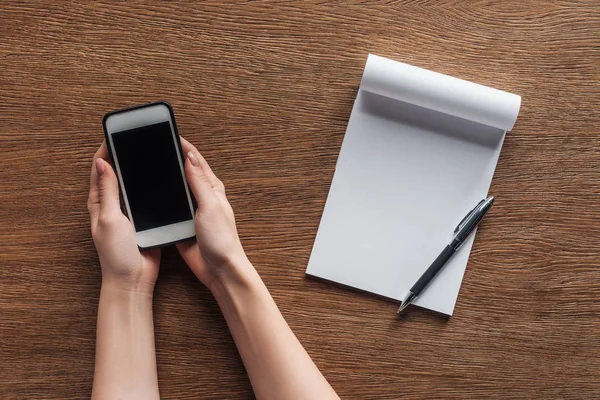 Vista parcial de la persona que sostiene el teléfono inteligente con la pantalla en blanco, pluma, cuaderno con espacio de copia sobre fondo de madera - foto de stock