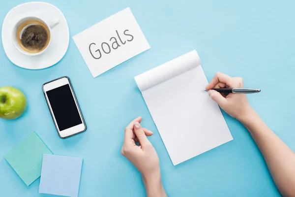 Cropped view of woman writing in notebook, lettering 'goals' on card, coffee, sticky notes and smartphone with blank screen on blue background — Stock Photo