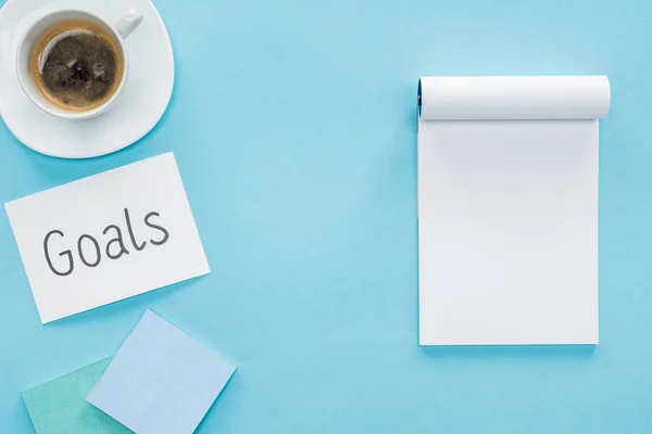 Vista superior de la tarjeta con letras 'metas', cuaderno en blanco y taza de café sobre fondo azul, concepto de fijación de metas - foto de stock