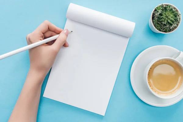 Vista cortada de pessoa escrevendo em caderno em branco com xícara de café no fundo azul — Fotografia de Stock