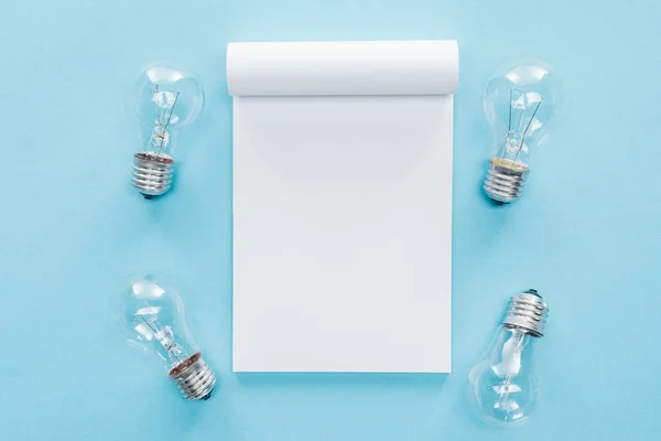 Vista superior del cuaderno en blanco con bombillas sobre fondo azul, lluvia de ideas y concepto de idea - foto de stock