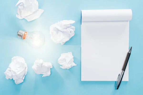 Top view of blank notebook with crumbled paper balls and glowing light bulb on blue background, having new ideas concept — Stock Photo