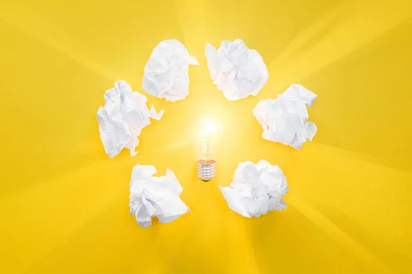 Top view of glowing light bulb in circle of crumbled paper balls on yellow background, having new ideas concept — Stock Photo
