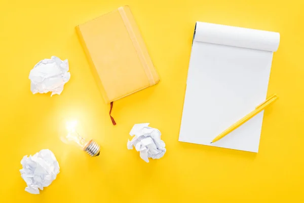 Vista superior del cuaderno en blanco, bolas de papel desmenuzadas y bombilla de luz brillante sobre fondo amarillo, con nuevo concepto de ideas - foto de stock