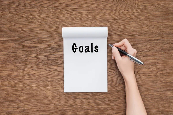 Cropped view of woman holding pen and notebook with 'goals' lettering on wooden background — Stock Photo