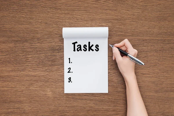 Vista recortada de la mujer escribiendo palabra 'tareas' en el cuaderno sobre fondo de madera - foto de stock