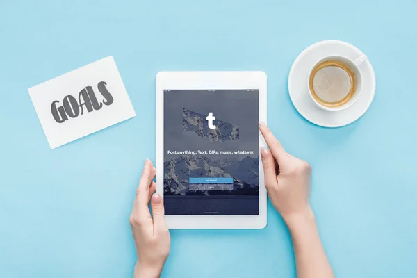 Corte vista da fêmea usando tablet digital com aplicativo tumblr na tela, metas lettering no cartão e café no fundo azul — Fotografia de Stock