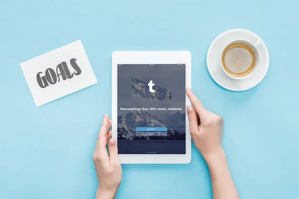 Vista recortada de la mujer usando tableta digital con aplicación tumblr en la pantalla, objetivos de letras en la tarjeta y café sobre fondo azul - foto de stock
