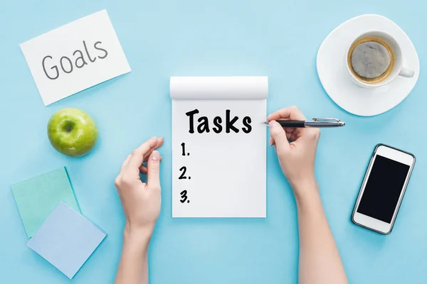 Vista recortada de la mujer escribiendo palabra 'tareas' en el cuaderno, letras 'objetivos' en la tarjeta, café y teléfono inteligente con pantalla en blanco sobre fondo azul - foto de stock