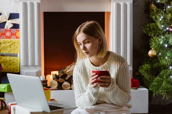 Junge Frau hält Tasse Kaffee in der Hand und benutzt Laptop in der Nähe von Kamin mit Brennholz — Stockfoto