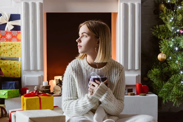 Mujer joven sentada, sosteniendo presente y mirando a la chimenea - foto de stock