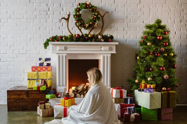 Young woman covered in blanket sitting with presents near fireplace with firewood at home — Stock Photo