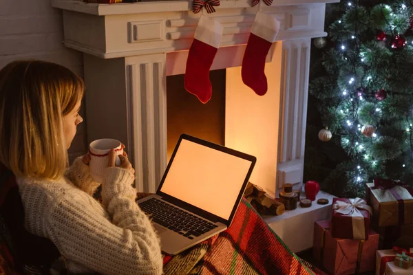 Giovane donna che tiene in mano una tazza di cacao e utilizza il computer portatile con schermo bianco nel periodo natalizio — Foto stock